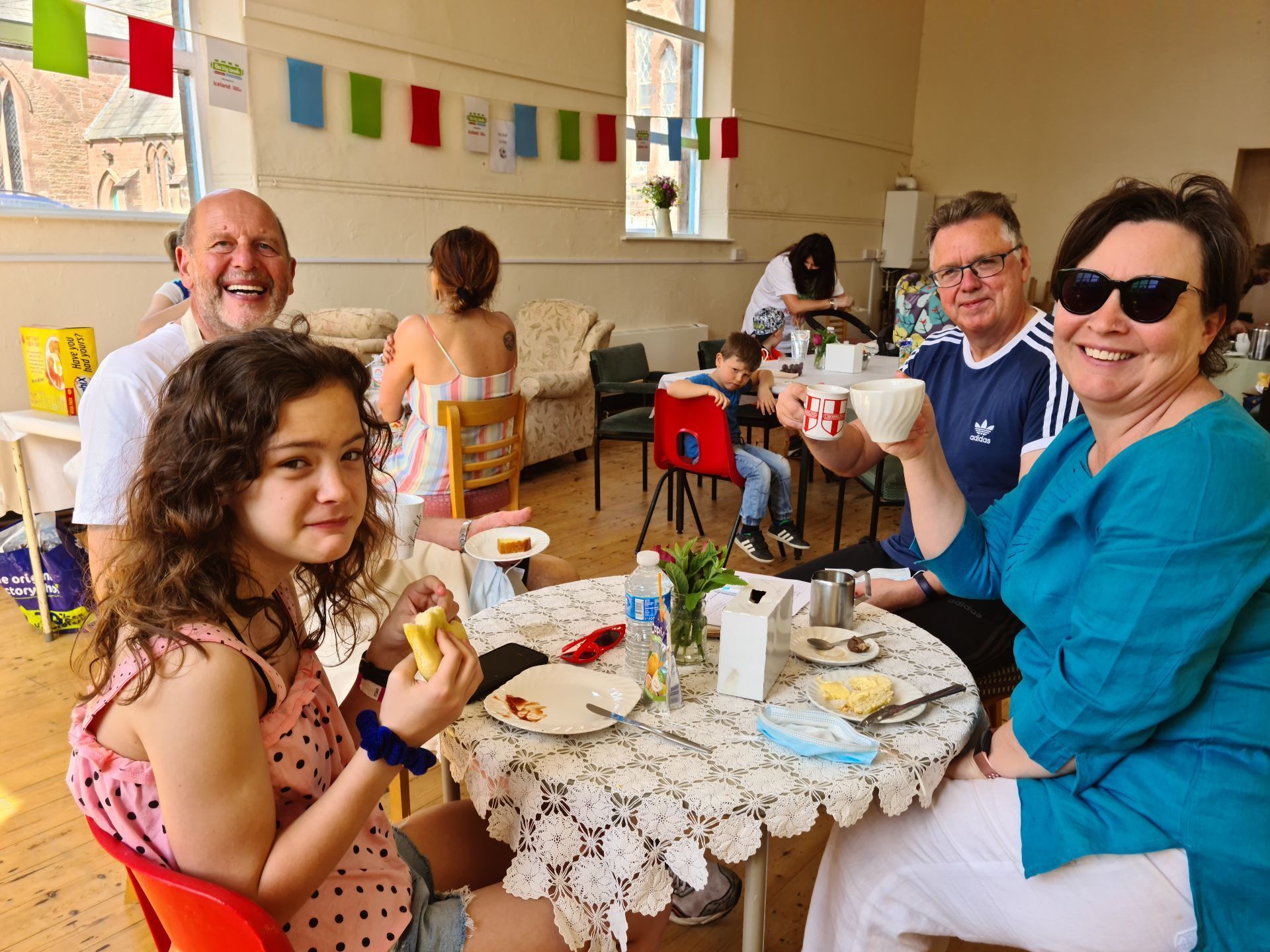 A group of people sitting at a table in a church hall, sharing drinks and food together. There is bunting up in the background and everyone is smiling