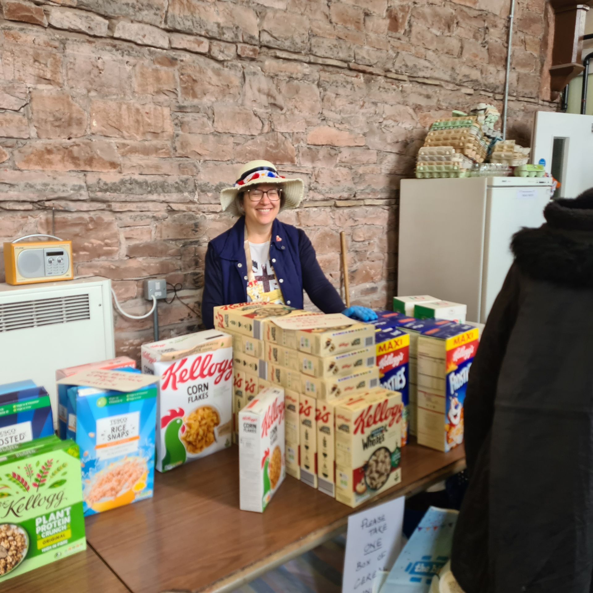 A volunteer at Wigton Food Pantry stands behind a table with boxes of cereal and other dried foods. She has a jazzy hat on and is smiling happily.