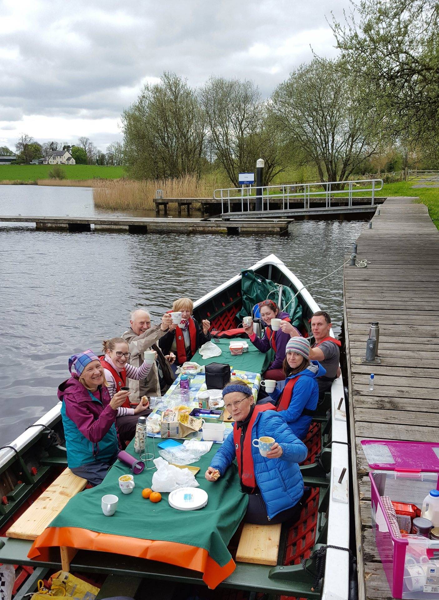 Group of people on a boat, sitting around a table sharing food and drinks. They are smiling at the camera and holding their mugs aloft.