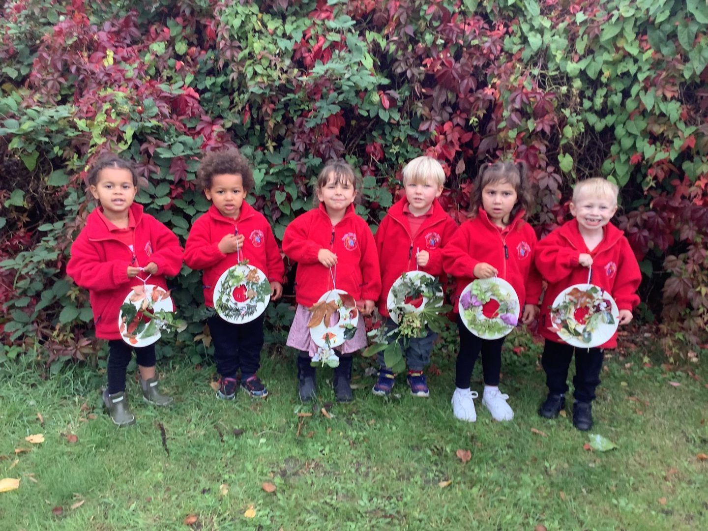Six very cute primary school children stand outside against a green and red maple tree. They are holding autumn wreaths they've created from leaves. They're ordered, boy, girl and are wearing red fleeces as part of their uniforms.