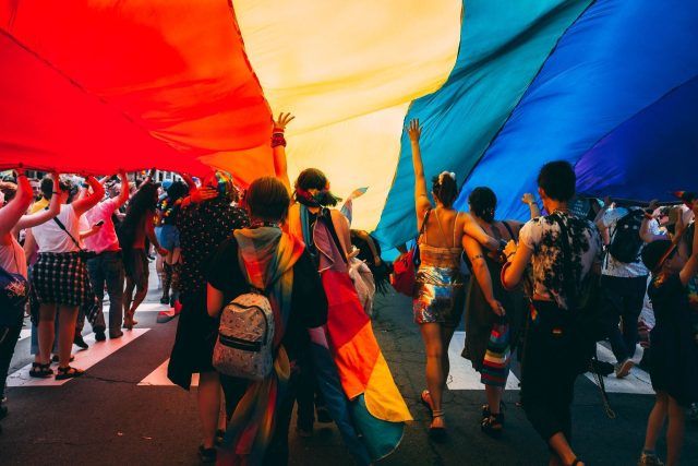 Group of people under a rainbow flag at a Pride event. You can only see their backs and the event looks vibrant and full of energy.