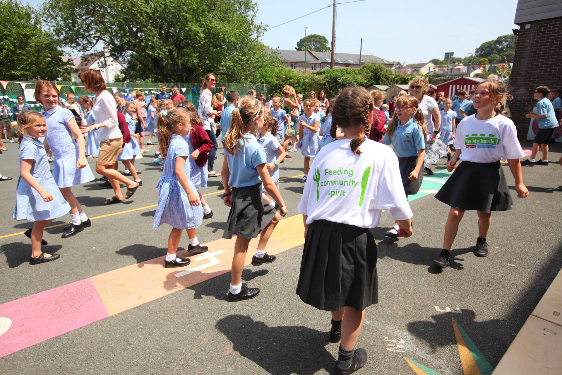Schoolchildren dancing in a playground. One is wearing a tshirt that says 'The Big Lunch' and one says 'Community spirit'