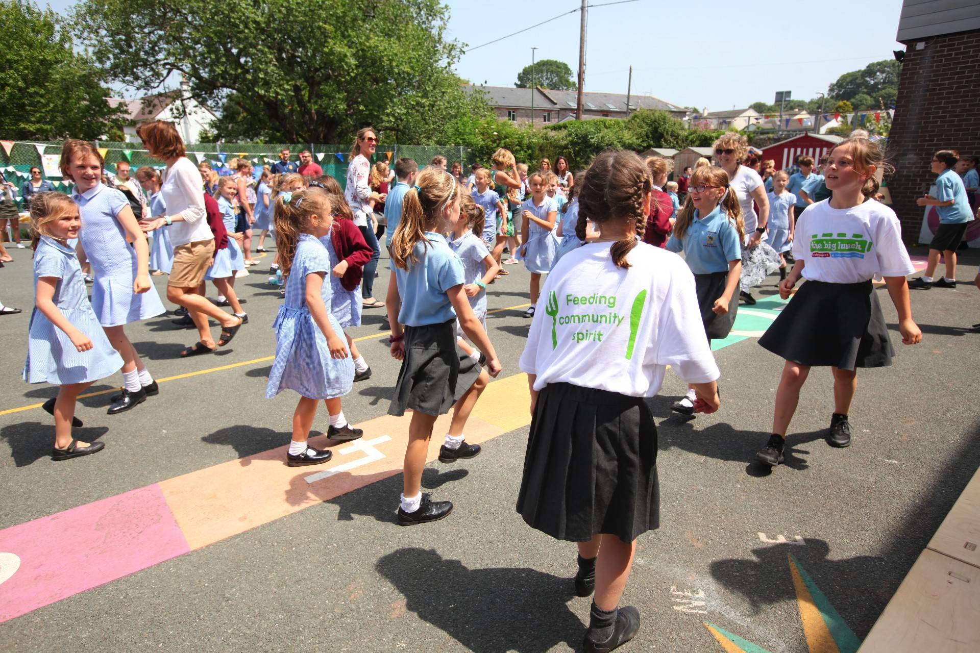 Plant ysgol yn dawnsio ar faes chwarae. Mae un yn gwisgo crys-t sy'n dweud 'Y Cinio Mawr' ac un yn dweud 'Ysbryd cymunedol'