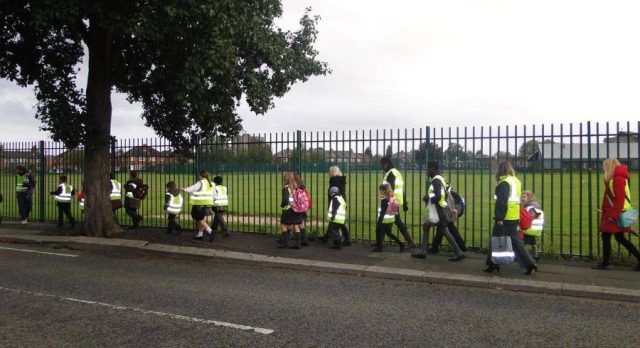 A group of children holding hands, wearing high-vis jackets, walking to school