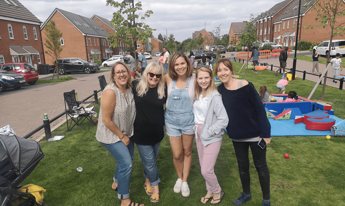 Five woman in green space surrounded by homes kids playing in the background