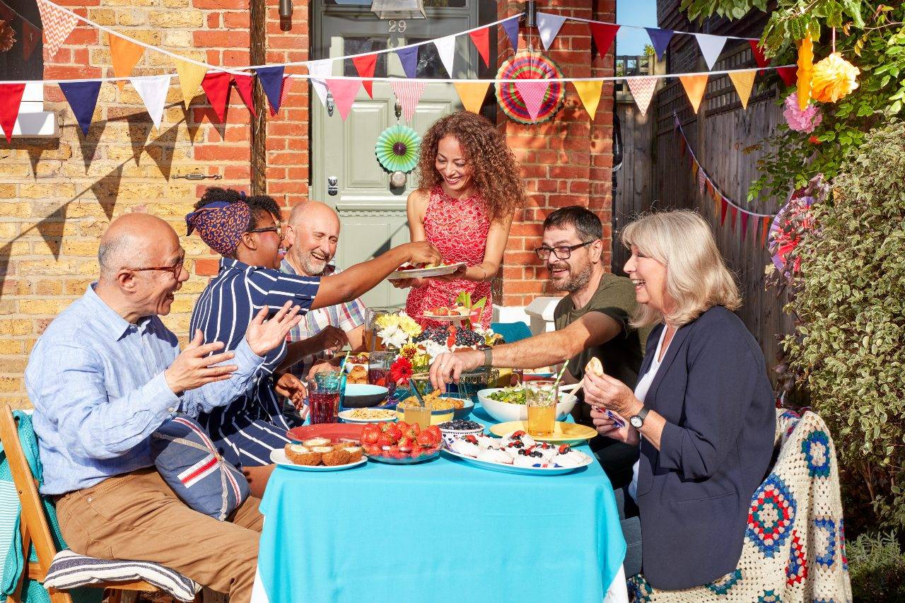 Group of people smiling and eating at a table outside a house. Bunting is strung across the scene and the sun is shining.