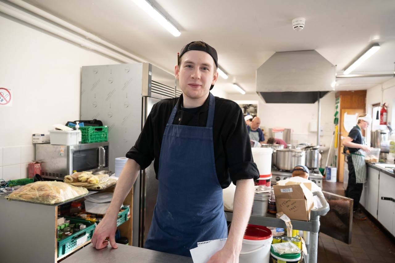 Man standing in commercial kitchen wearing apron and smiling at the camera.