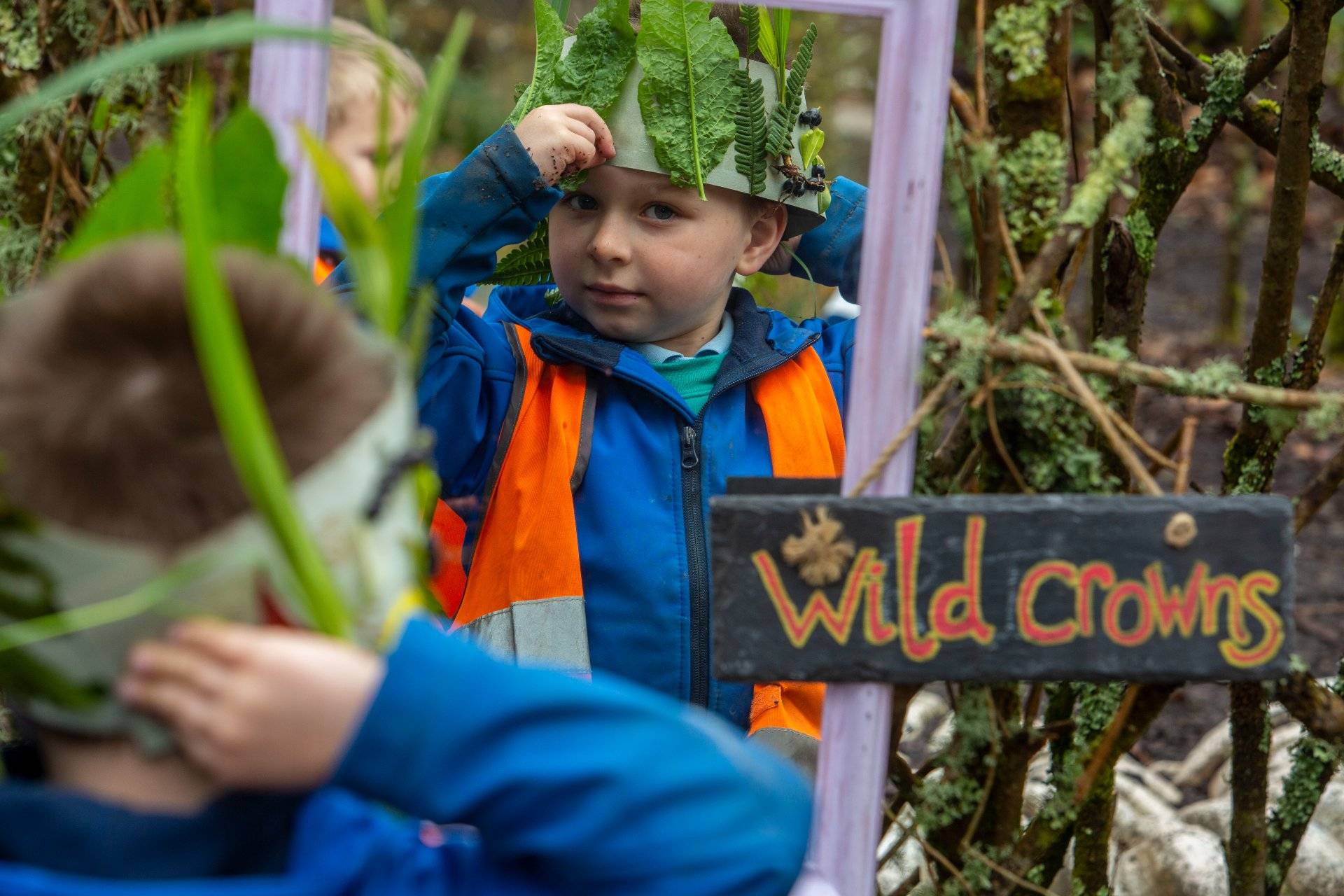 Bachgen bach yn gwisgo hwdi glas a high-vis oren yn rhoi coron wedi'i gwneud o ddail ymlaen, yn sefyll wrth ymyl arwydd yn dweud 'Coronau gwyllt'