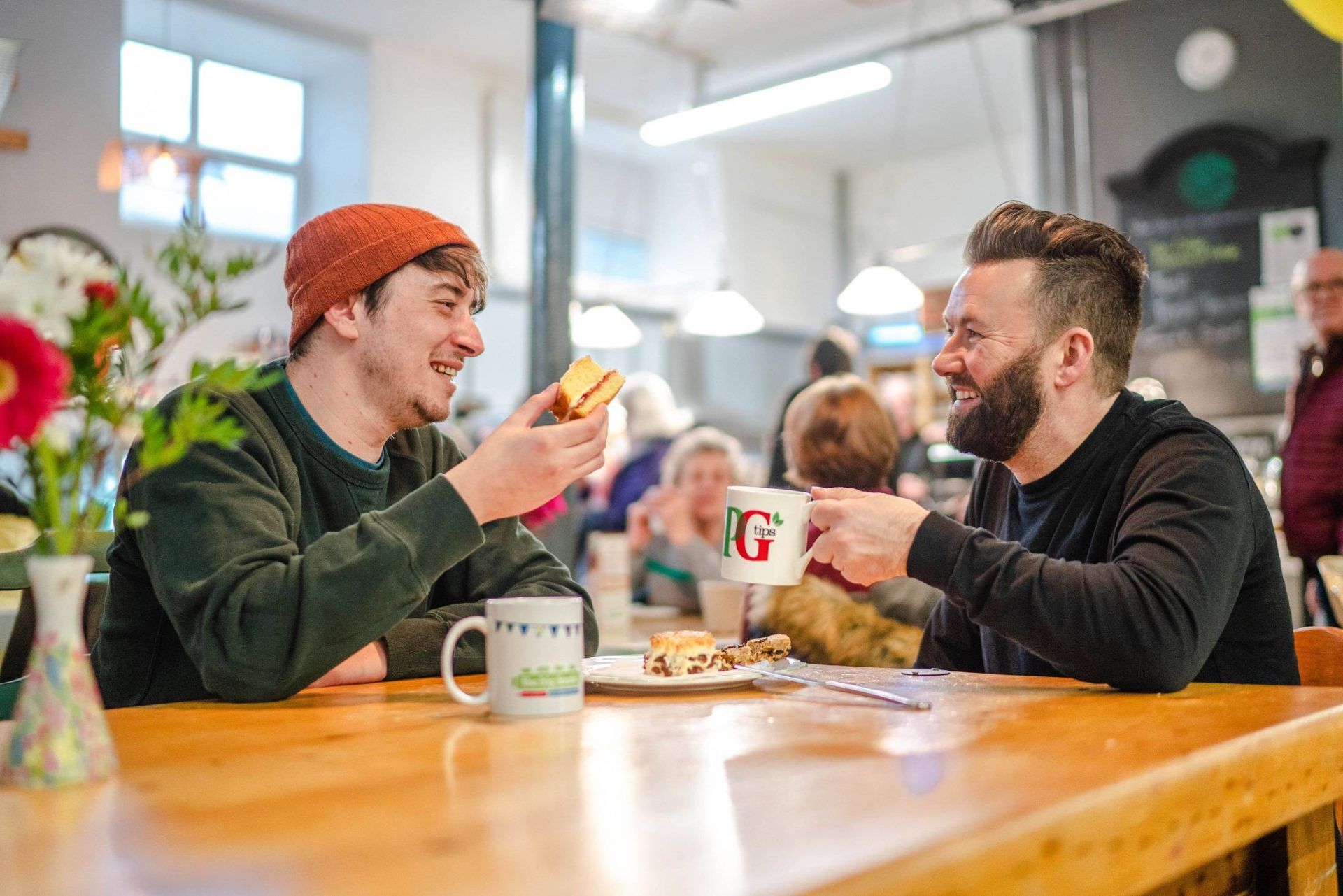 Two men laughing and enjoying a cup of tea at an indoor event.