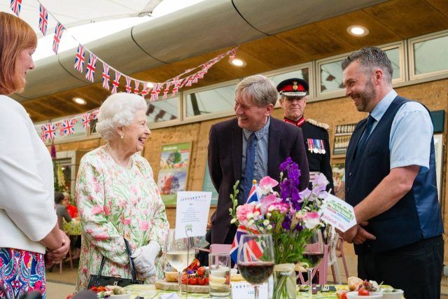 Queen Elizabeth II smiling and chatting with Peter Stewart from the Eden Project Communities team. There is food and drink laid out on a table in the foreground, and Union Jack bunting behind.