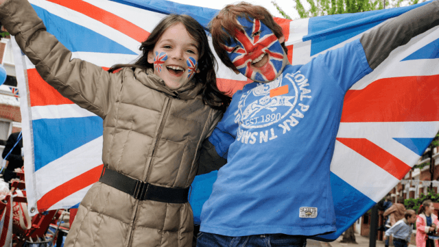 Two children draped in a Union Jack flag smiling at the camera. One has their face painted.