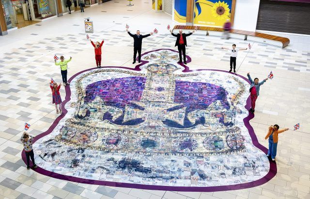 A group of people standing around a piece of art waving Union Jack flags. The art is a Coronation Crown made up of lots of photographs laid on the floor.