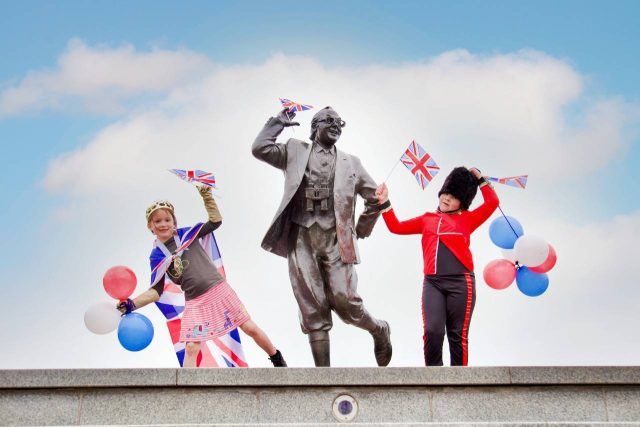 Two children post with their arms up next to the dancing Eric Morecambe statue. They're holding red, white and blue balloons and holding Union Jack flags, as part of the Coronation Big Lunch celebrations.