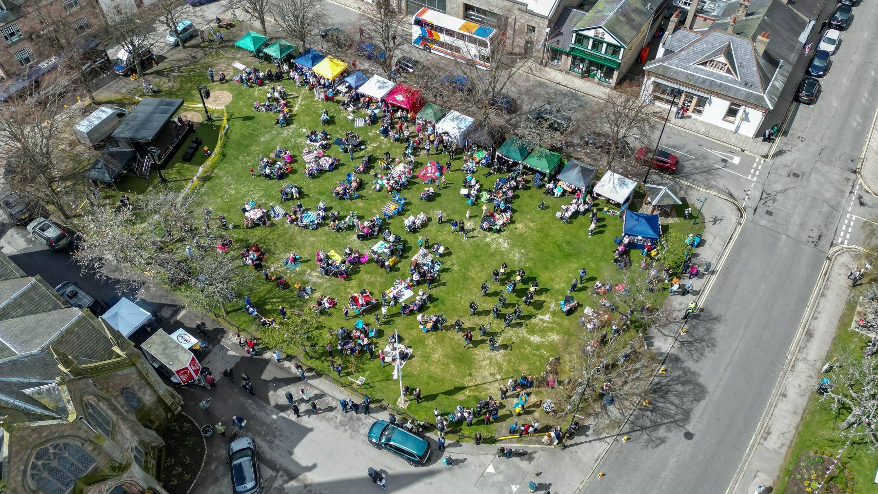 Drone footage of the Ballater Coronation Big Lunch - groups of people sitting around picnic tables and a band performing can be seen. Tents line one side of the green space.