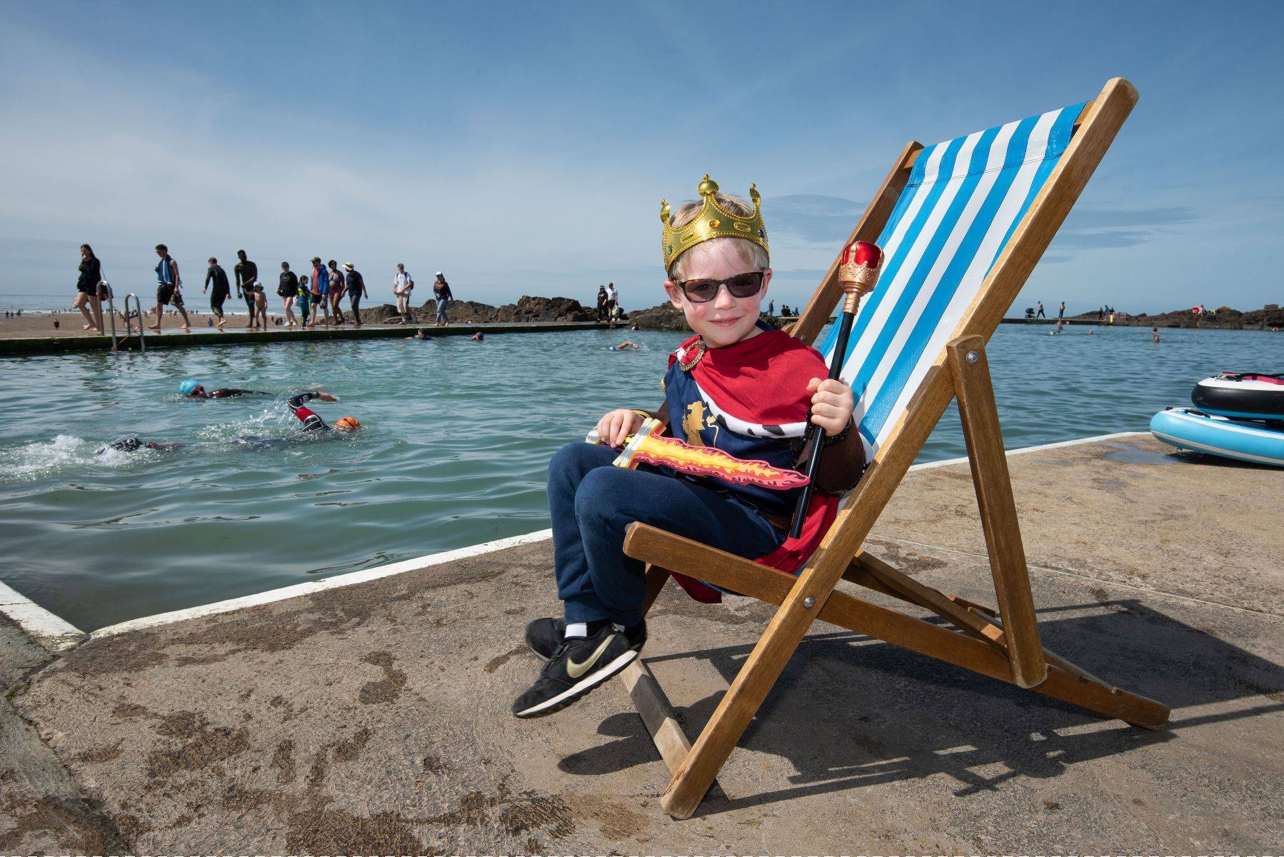 A small boy sits in a big deckchair wearing sunglasses and a paper crown. Bude Sea Pool is visible in the background.