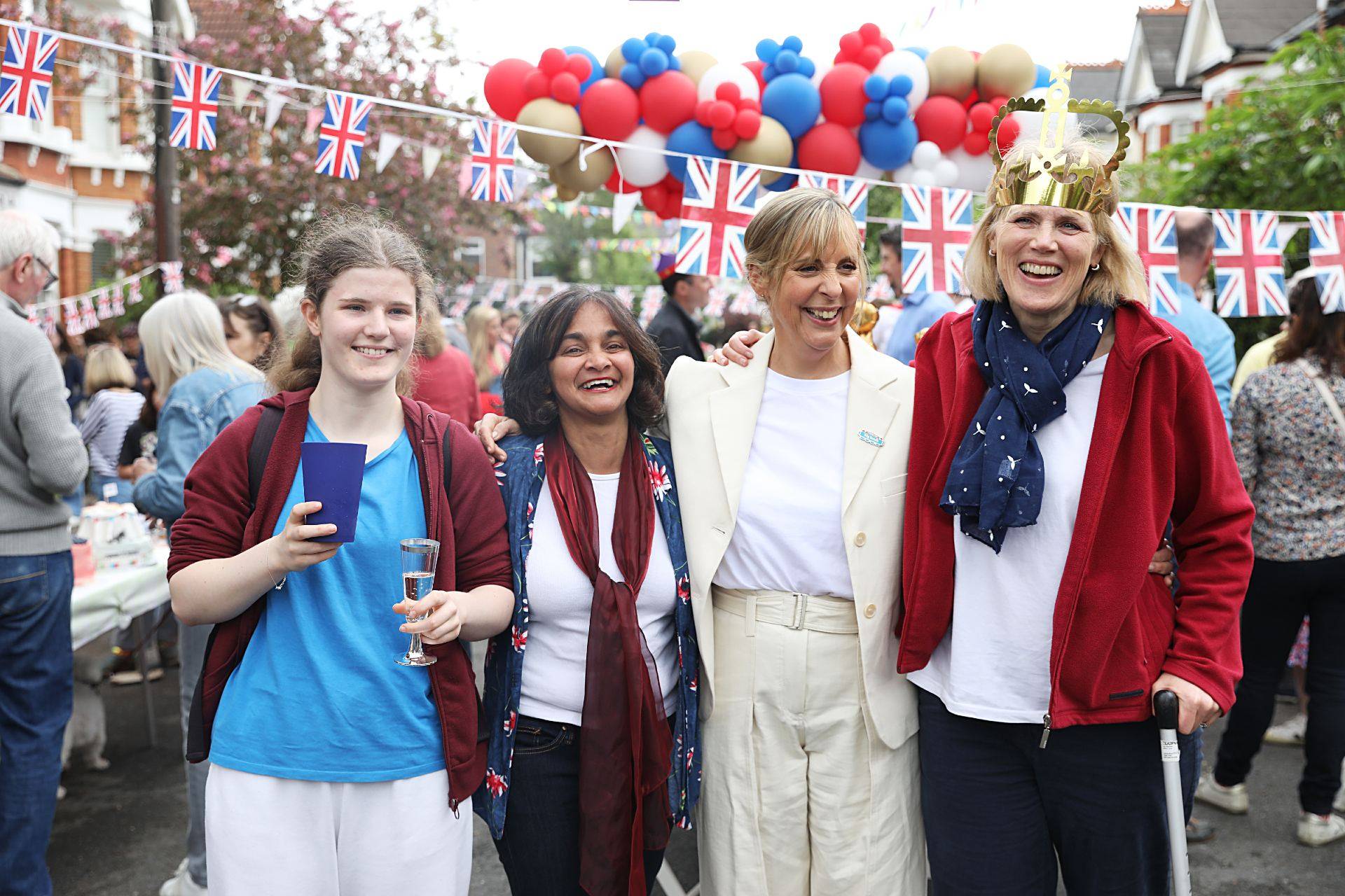 Mel Giedroyc celebrates with residents of Grosvenor Gardens. She has her arm around two women and they're smiling, with red white and blue balloons and Union Jack bunting in the background. The scene behind them is bustling with people.