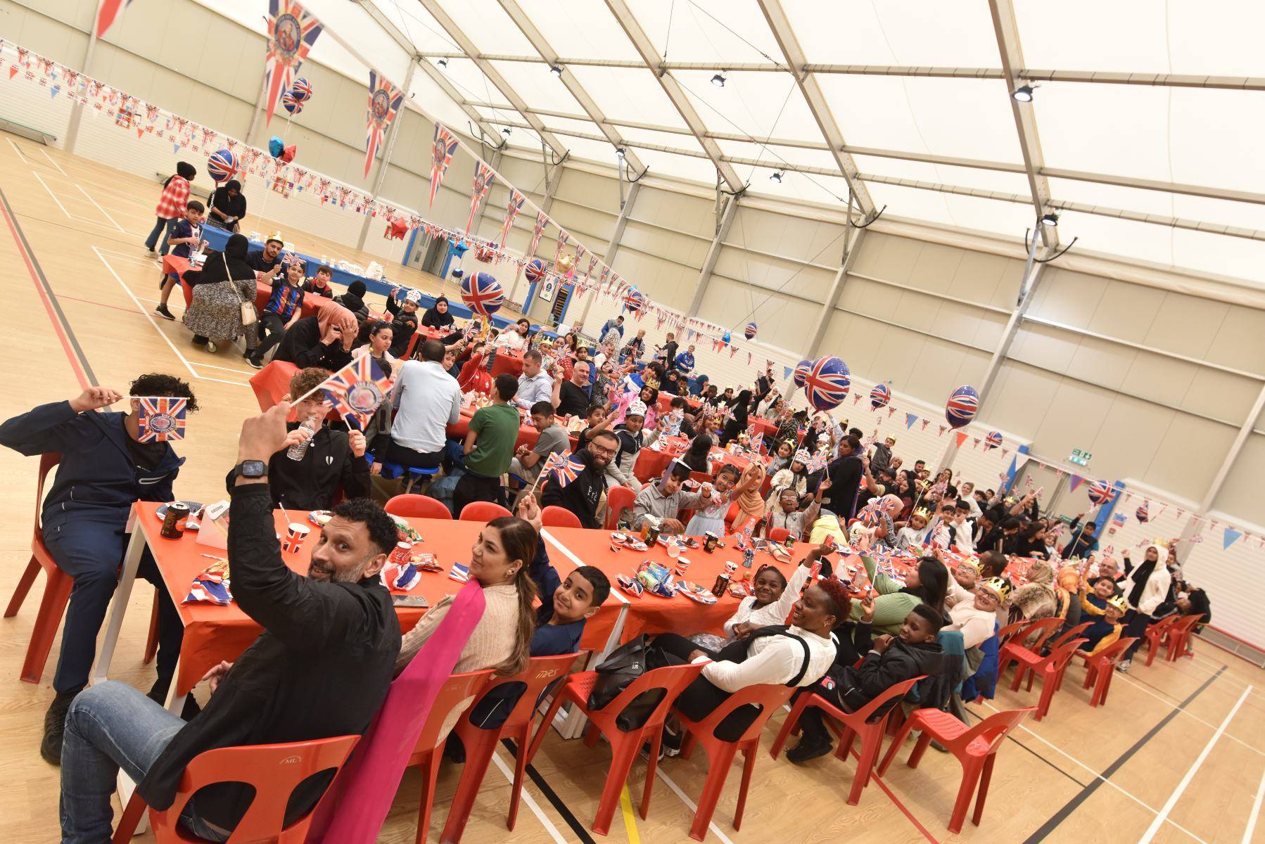 A large group of people sit around tables and chairs in the sports hall and smile up at the camera