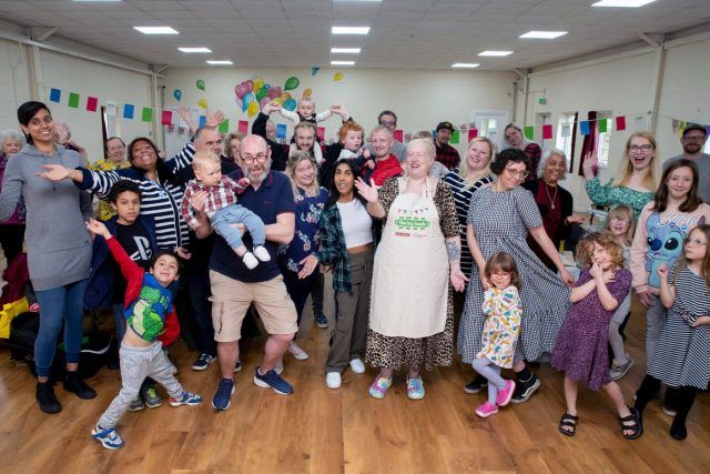 Lucy Scott, who runs Lil's Food Parlour, wearing a Big Lunch apron, surrounded by a big group of cheering people, in West Heath community centre.
