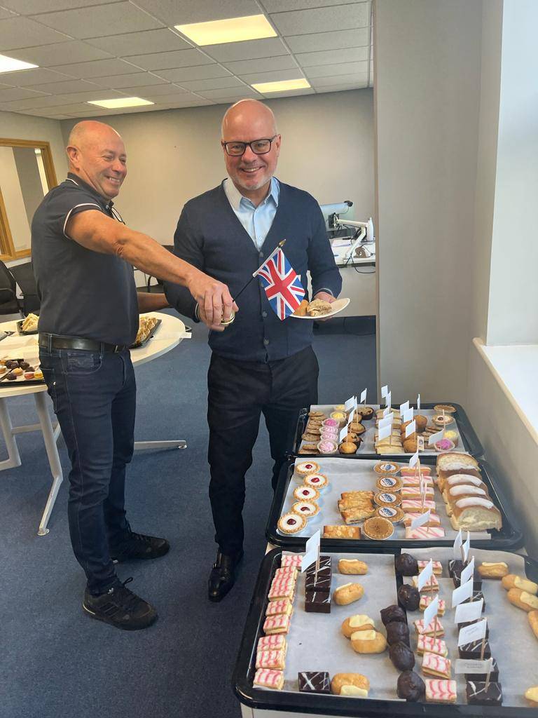 Two men shake hands in an office - one is holding a Union Jack. There is a tray of cakes in the foreground.