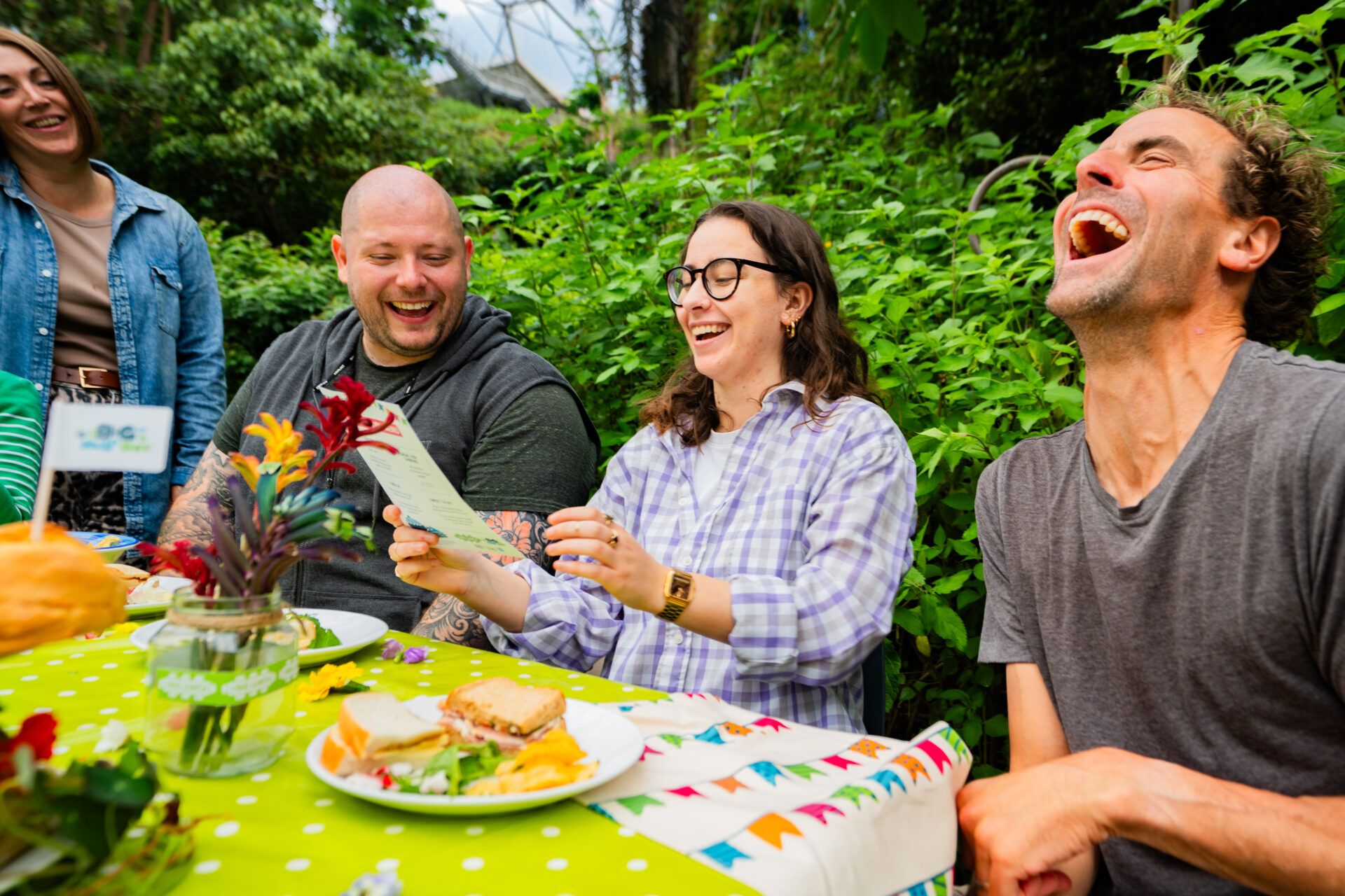 People sit laughing at a table, looking at a conversation menu. There are cakes in front of them and greenery visible behind. The atmosphere looks relaxed.