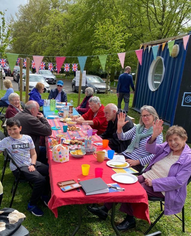 People sat at a long table with a red tablecloth under colourful bunting. They are smiling at the camera, holding their drinks up and generally enjoying themselves.