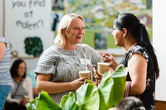 Two woman chatting at an event. They're holding hot drinks and wearing name badges, and look engaged in their conversation.