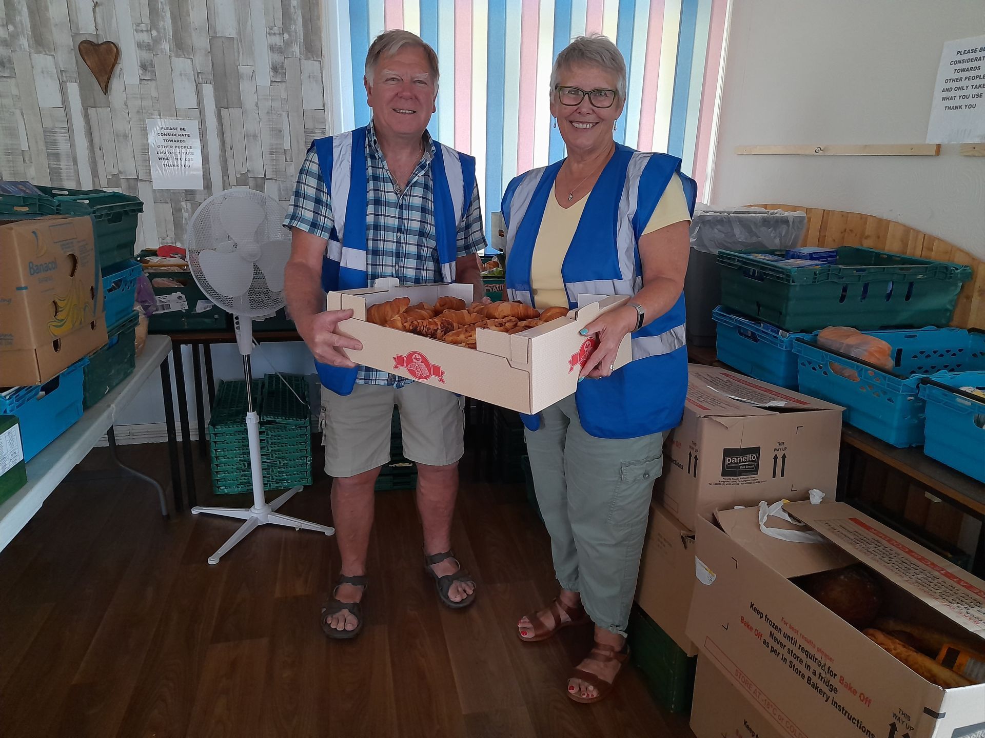 Two volunteers wearing blue hi-vis vests are holding a box of produce between and smiling. There are more boxes ready to unpack in the background.