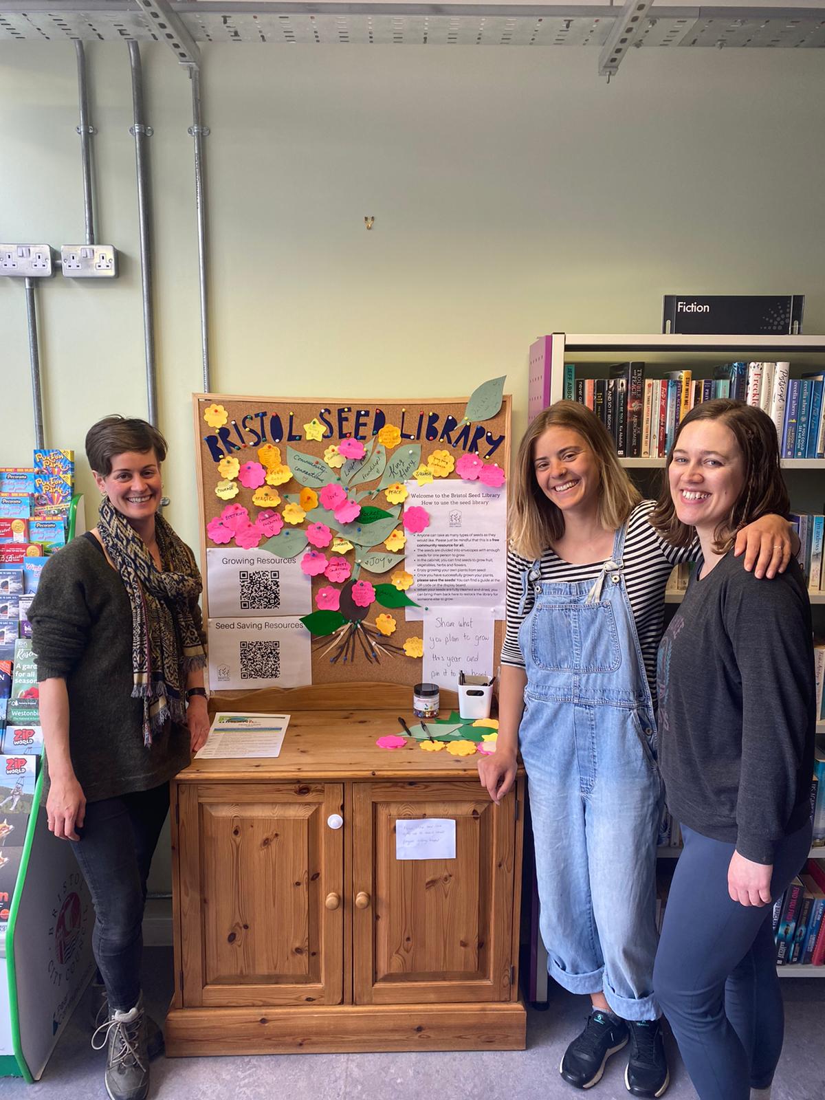 The three founders of Bristol Seed Library standing in front of the dresser containing their seeds.