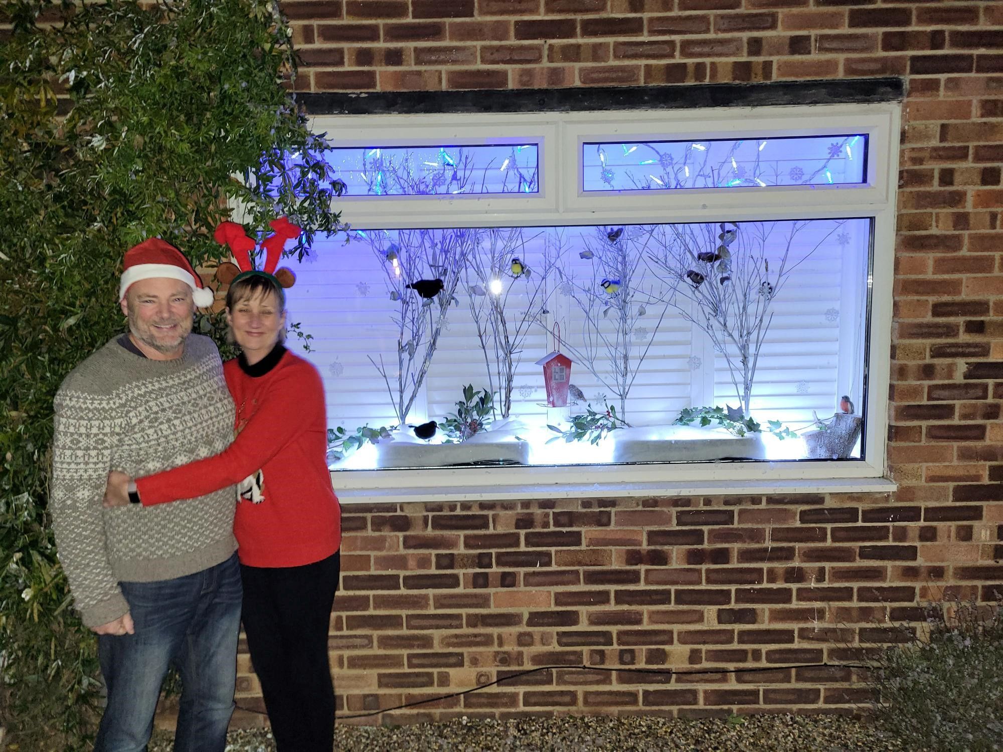 Angie and her partner are wearing Christmas jumpers and standing outside their advent window, which shows winter trees and birds.