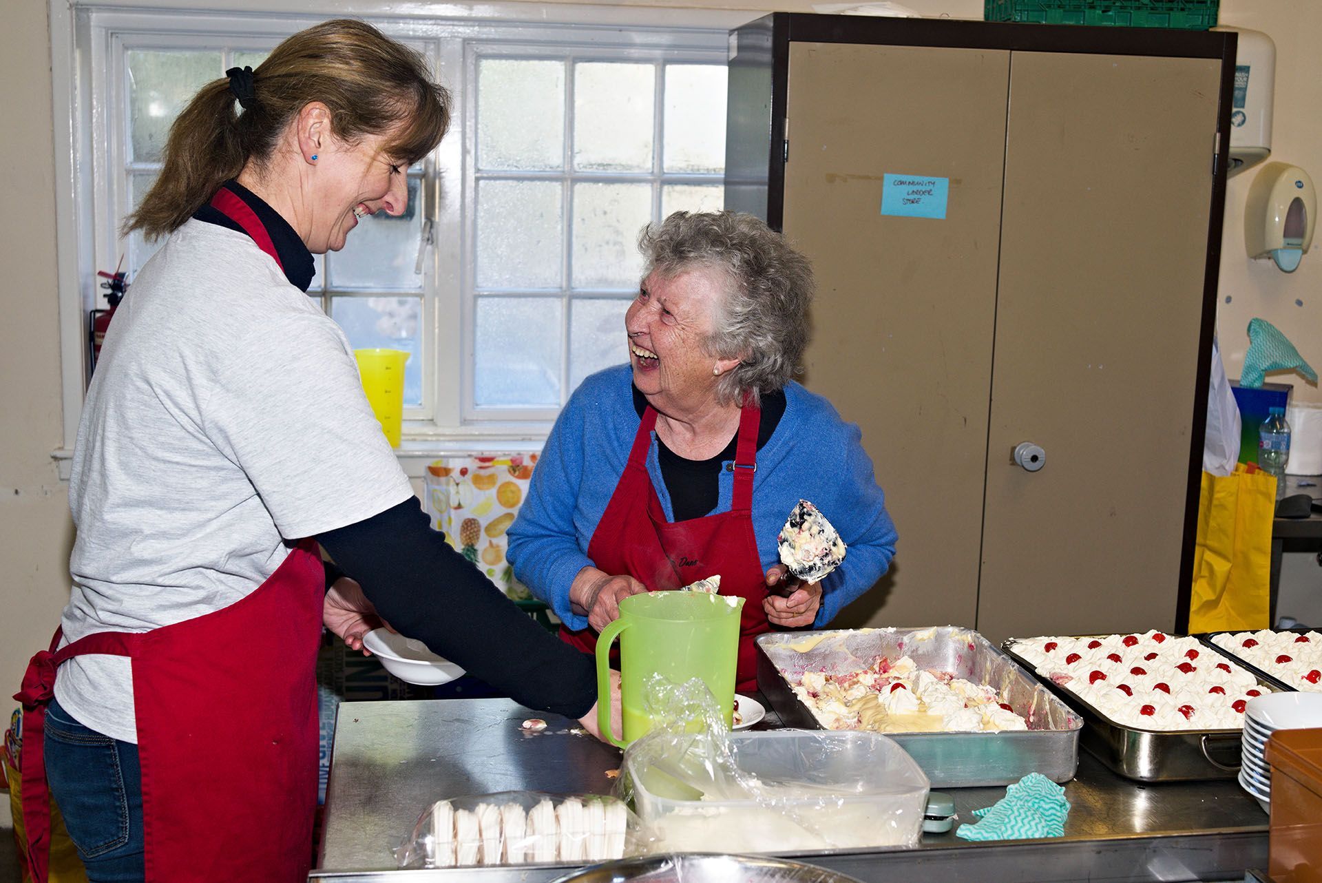 Two women at a Big Lunch at Christmas prepare trifle for the community. They're laughing and look full of Christmas spirit.