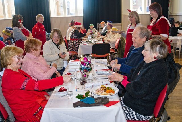 A group of elderly people sit at a table pulling crackers for their Christmas lunch. They're smiling and laughing, and other tables are visible in the background.