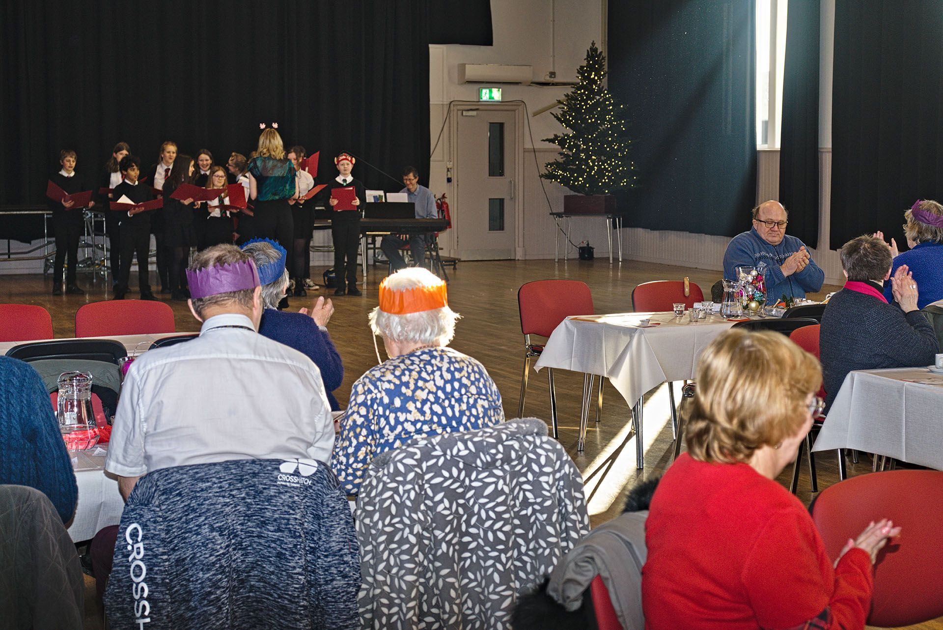 A group of people watching some children singing carols in a village hall. They're smiling and clapping.