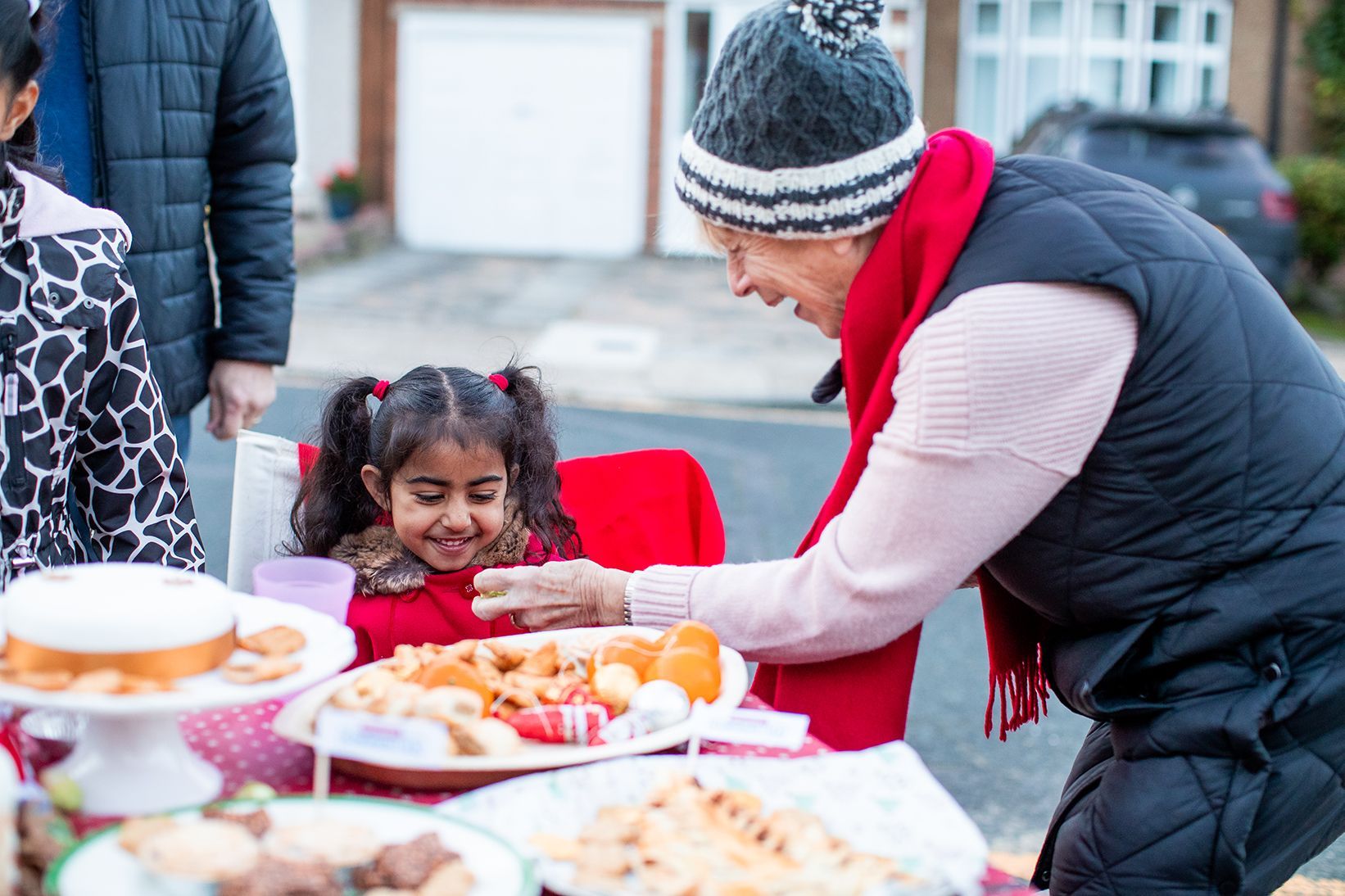 An older woman gives a young girl a mince pie - they're both smiling and wrapped up warm. You can see a table full of delicious festive food.