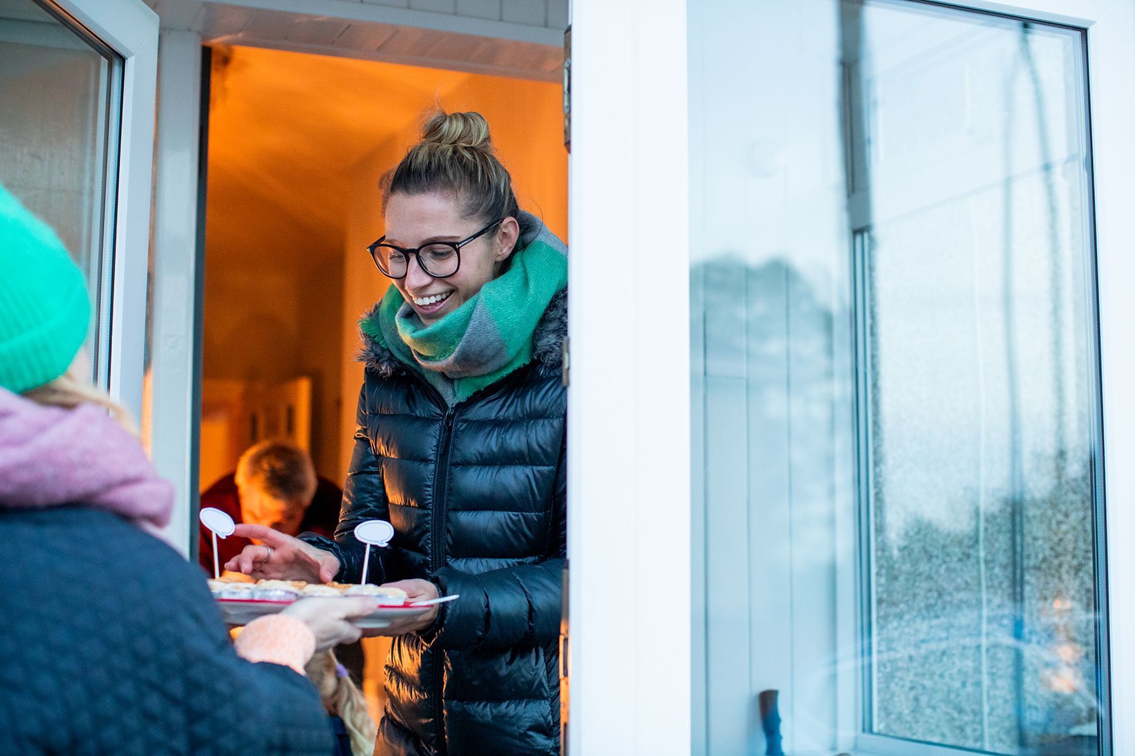 A woman answers the door smiling, as the person who knocked holds up a plate of mince pies.