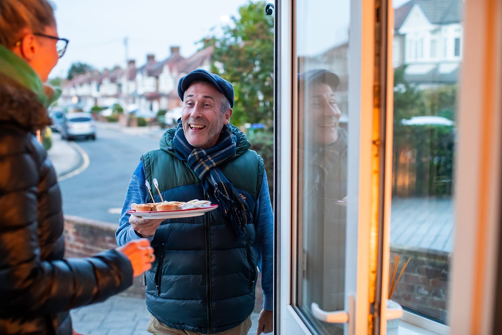 A man stands on a doorstep proffering a plate of mince pies. He's got a huge smile on his face.