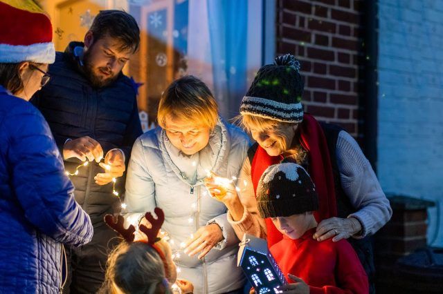 Group of people hanging fairy lights outside. Two women reach down towards children and everyone is smiling. The photo is full of warmth and cheer.