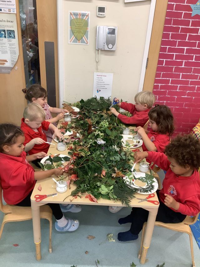 Children sitting around a table which has foliage and leaves on top - they're sticking leaves onto a circle of paper.
