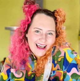 Biography picture of Bake Off's Lizzie Acker. Lizzie is smiling and her hair is coloured pink on one side and orange on the other. She is wearing a colourful printed jacket.