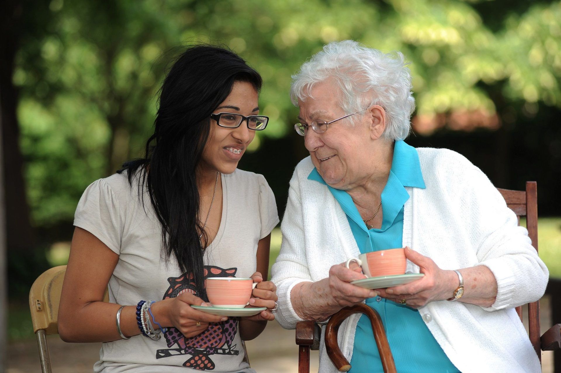 Two ladies sit outside with cups of tea and share a joke. One of them is in her twenties, the other is in her seventies.