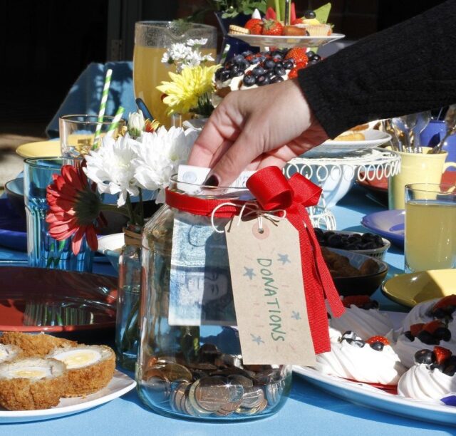 A glass donation jar is having a 5 pound note added amidst a beautifully set table containing cakes, drinks and flowers.