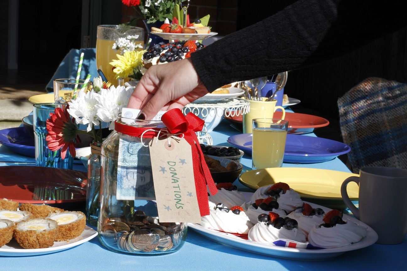 A glass donation jar is having a 5 pound note added amidst a beautifully set table containing cakes, drinks and flowers.