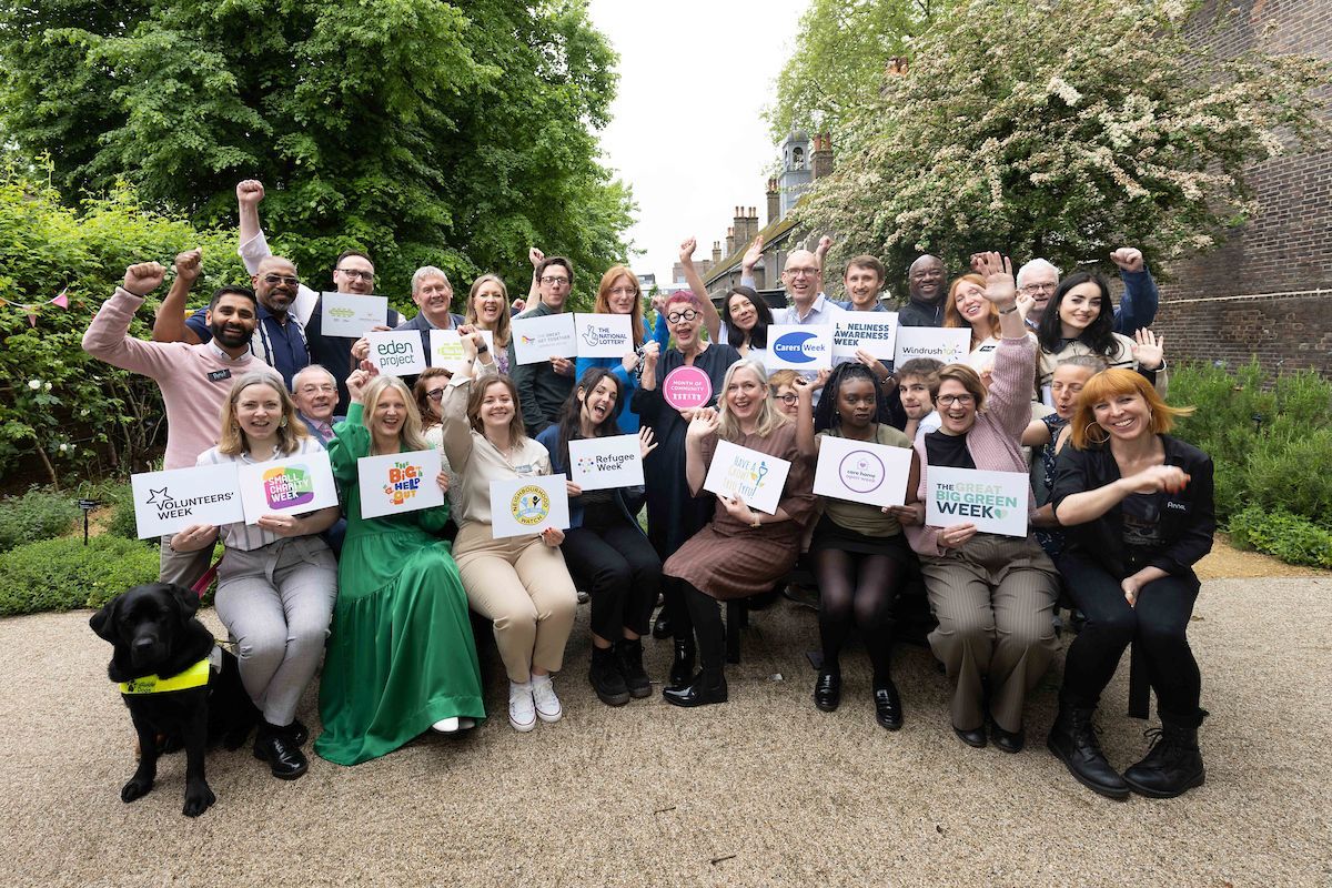 Month of Community partners, all standing together in a group outside and smiling at the camera. They're holding up signs with the names of their organisations.