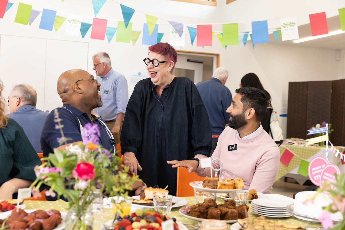 Jo Brand chatting to Month of Community partners at a Big Lunch event. There is a table full of delicious food and bunting strung up in the background.