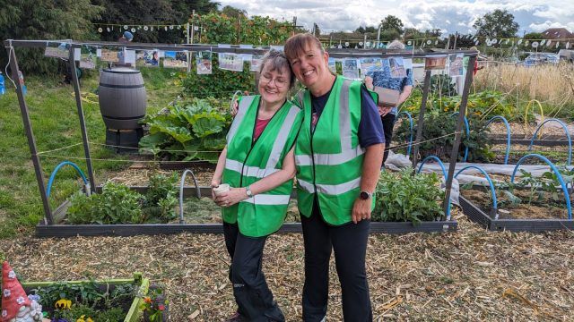 Tracey and a volunteer wear green high vis jackets at their community allotment. Plants are visible in the background