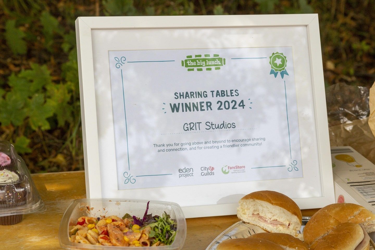 A framed certificate announcing that GRIT Studios are the Sharing Tables Winner 2024. The certificate sits on the Big Lunch table in GRIT's community garden.