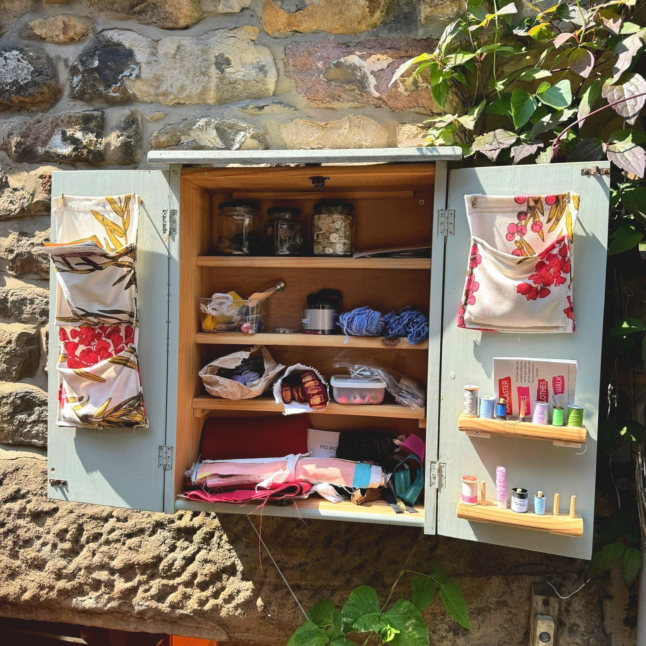 A small cupboard reminiscent of a spice cupboard is attached to a stone wall in Lancaster. The cupboard is full of jars of buttons, threads and other sewing items. The sewing cupboard is painted pale blue and looks lovely in the sunshine.