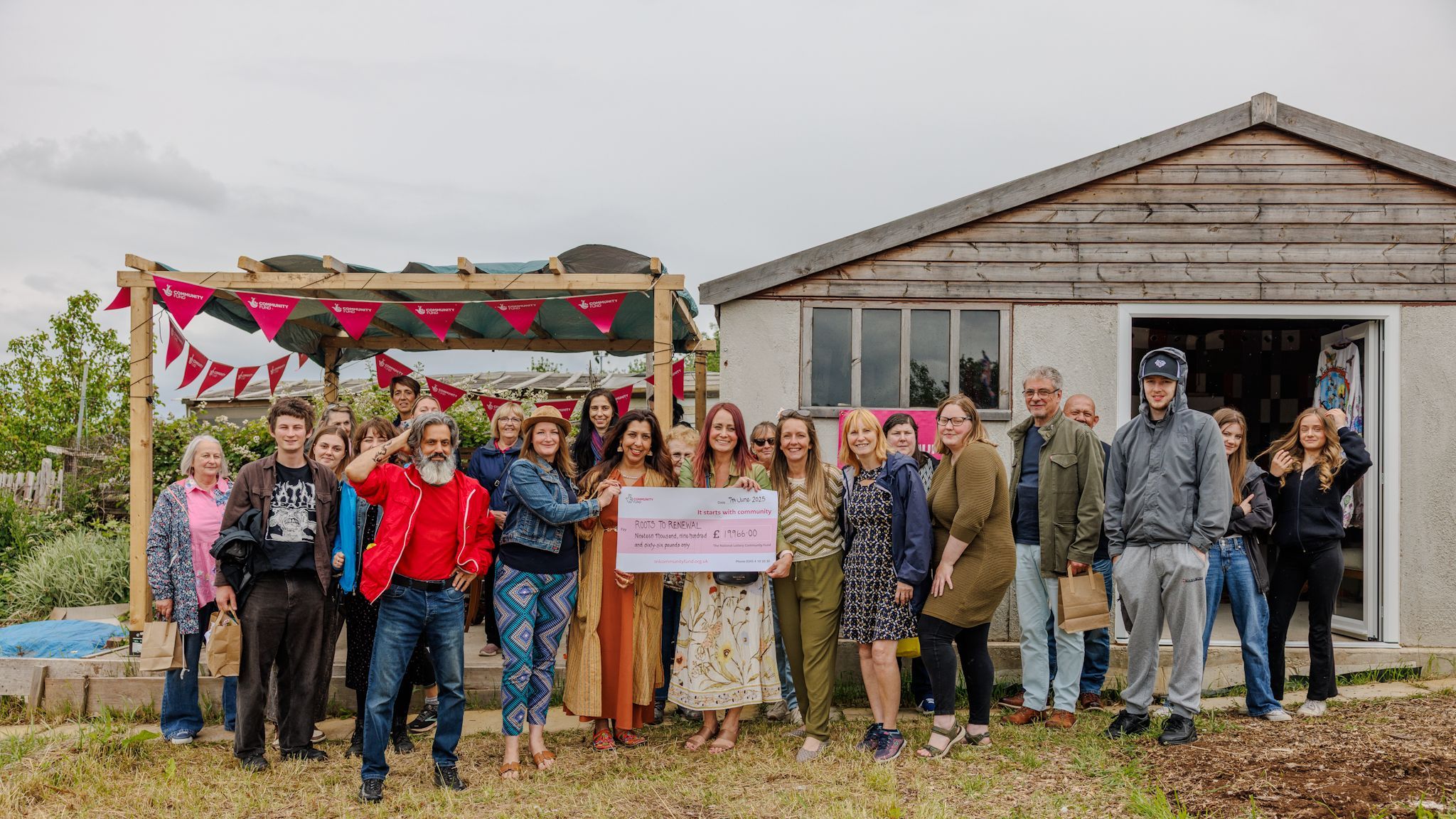 Roots to Renewal CIC Big Lunch opening party. A group of people standing in front of a wooden shed, holding a National Lottery cheque aloft.