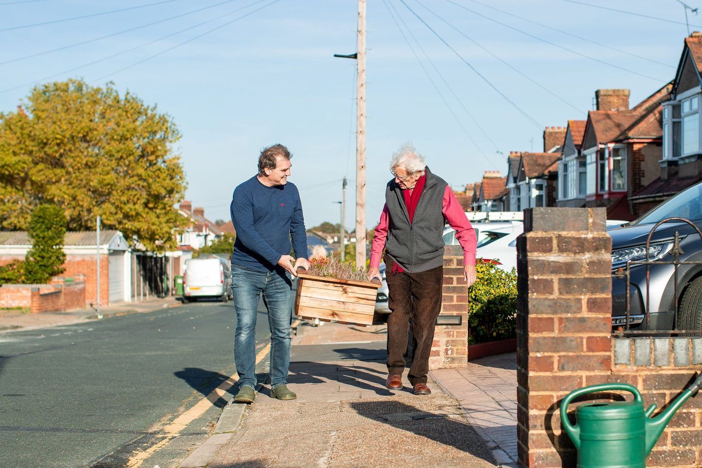 Two male neighbours carrying a planter along the street to one of their houses. They're helping one another, sharing resources and chatting. The sun is shining and they look friendly and happy.