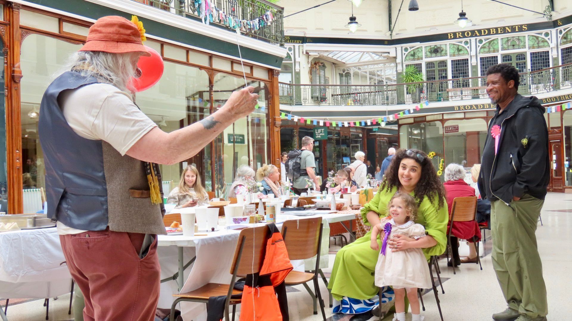 A man wearing a waistcoat and orange hat blows bubbles for a delighted little girl
