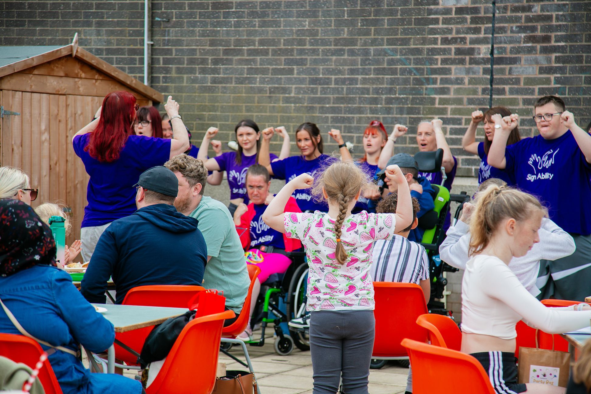 A group of people in purple tshirts are standing in a line against a wall and performing in a Makaton choir to the community sitting at their tables at the Love Barrow Libraries Big Lunch. A young girl in the crowd is joining in and copying the choir's Makaton gesture.